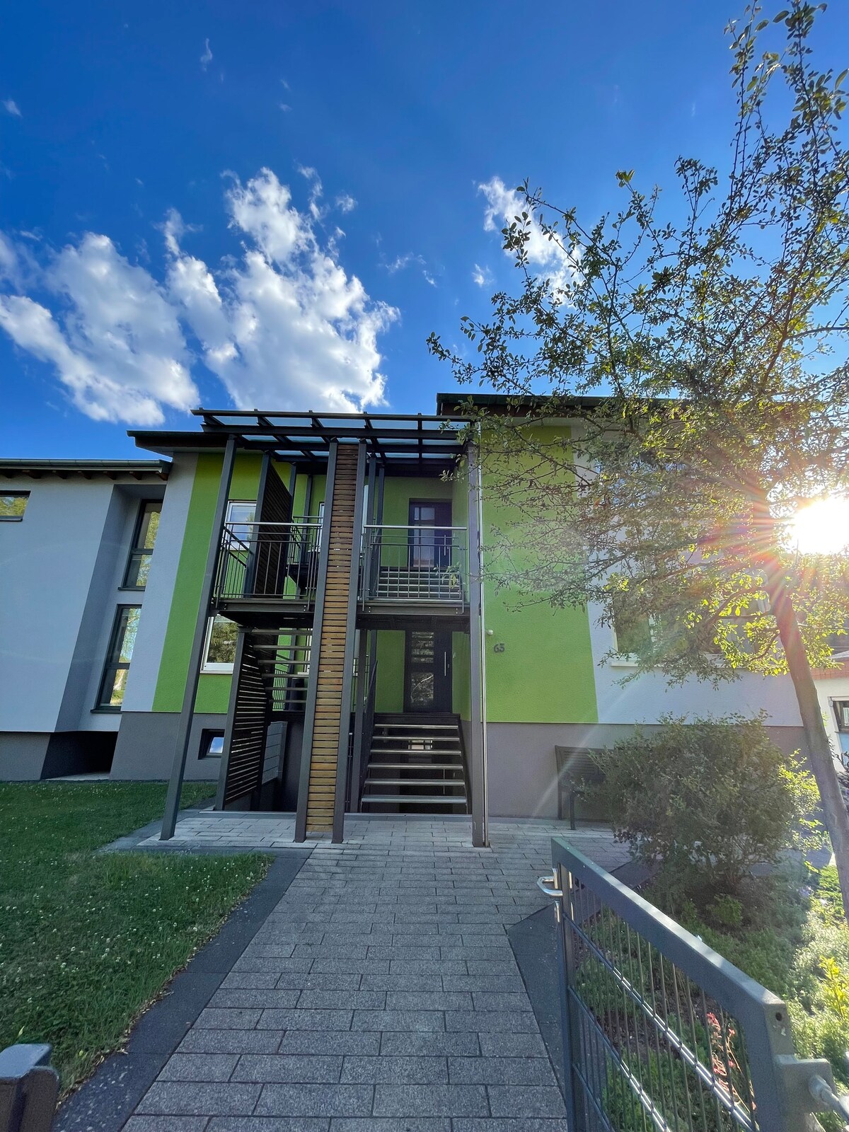 The exterior of the vacation rental showcases a modern two-story building featuring a combination of green and neutral-toned walls. A well-maintained pathway leads to the entrance, flanked by grass and trees. Natural light shines down from a clear blue sky, enhancing the inviting ambiance.