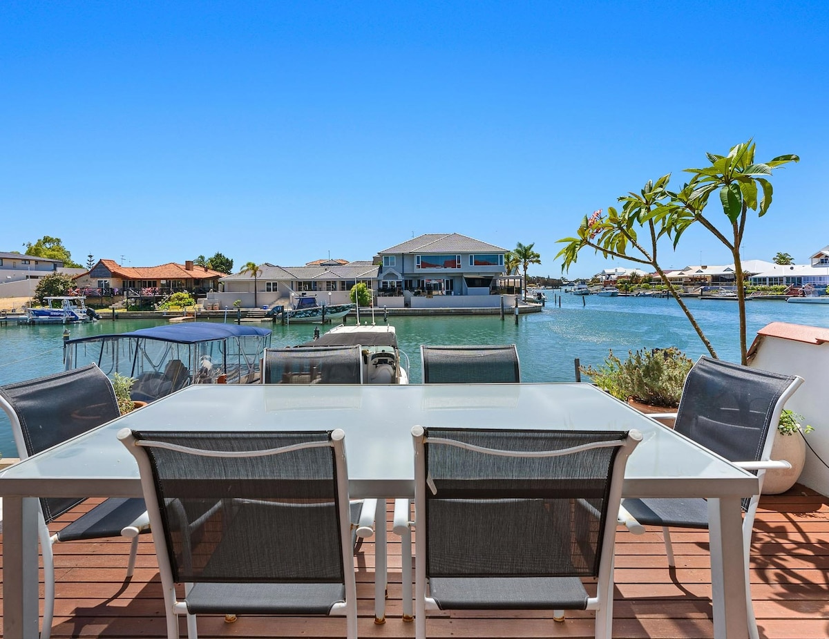 A spacious outdoor dining area features a glass table surrounded by six lightweight chairs, overlooking the tranquil waters and neighboring villas. Lush greenery adds a touch of nature to the setting, enhancing the inviting waterfront ambiance.