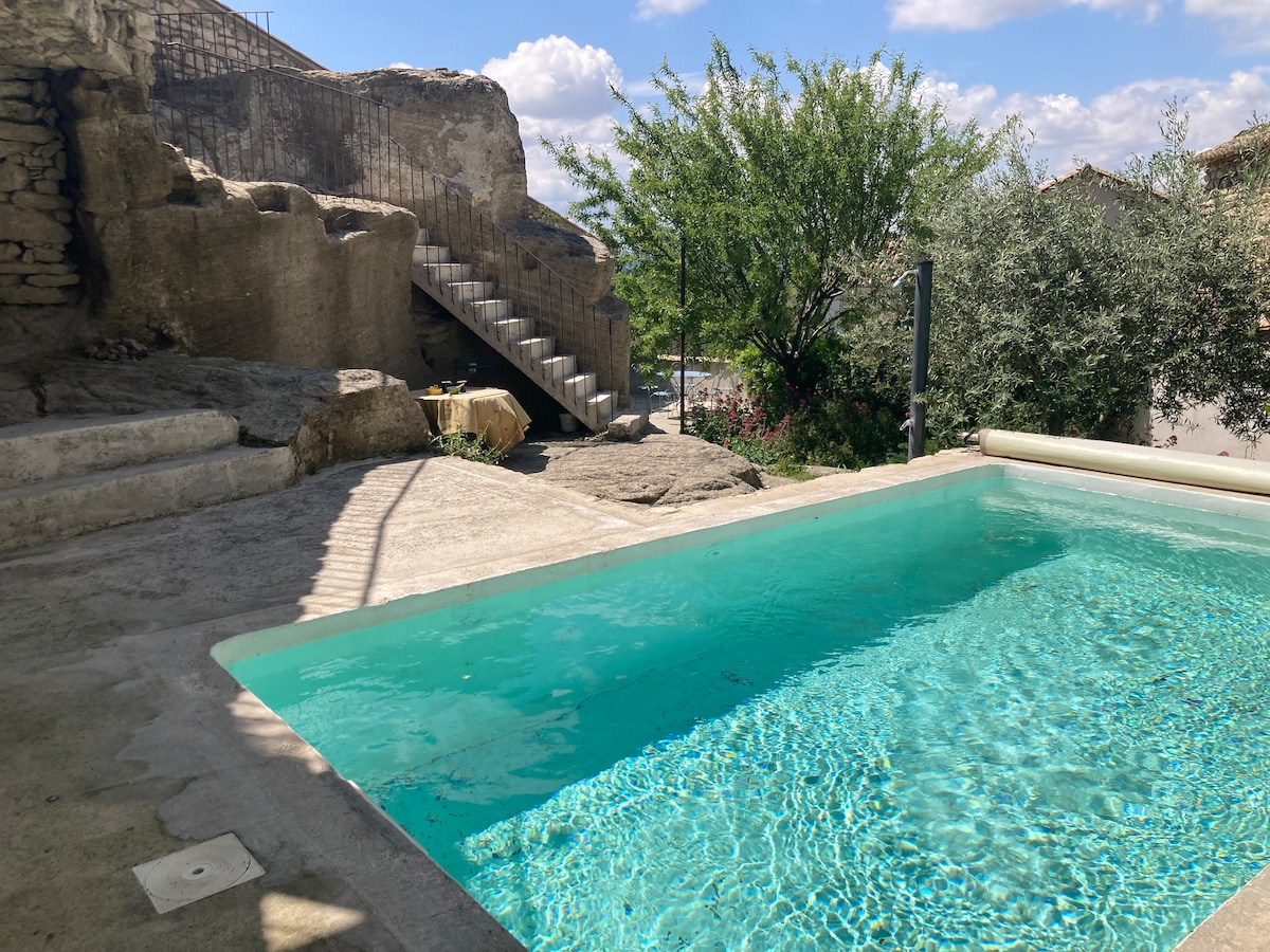 A serene pool area is surrounded by natural rock formations and greenery. Steps lead to a stone pathway, while the water features a clear blue hue. Sunlight casts gentle shadows on the textured concrete deck, providing a tranquil outdoor space.