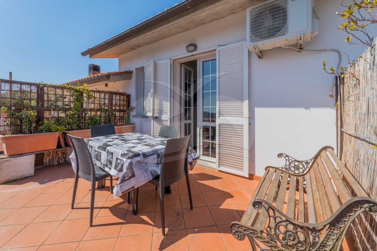 A spacious terrace is equipped with a table and chairs, offering a comfortable outdoor dining space. Lush greenery is visible in decorative planters, and sunlight brightens the area. A railing provides privacy, while an air conditioning unit is mounted on the wall.