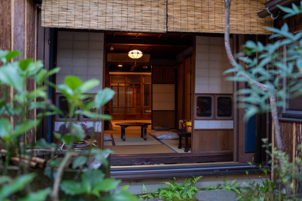 An entrance reveals a traditional interior, featuring tatami flooring and wooden paneling. A low wooden table is positioned at the center, surrounded by floor cushions. Natural light filters through the sliding doors, while greenery from the garden provides a serene backdrop.