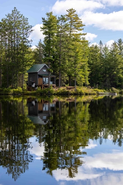 A cabin sits serenely at the edge of a private lake, surrounded by lush green trees. The water reflects the structure and natural scenery, creating a peaceful atmosphere. The clear sky above features soft clouds, enhancing the tranquil setting.