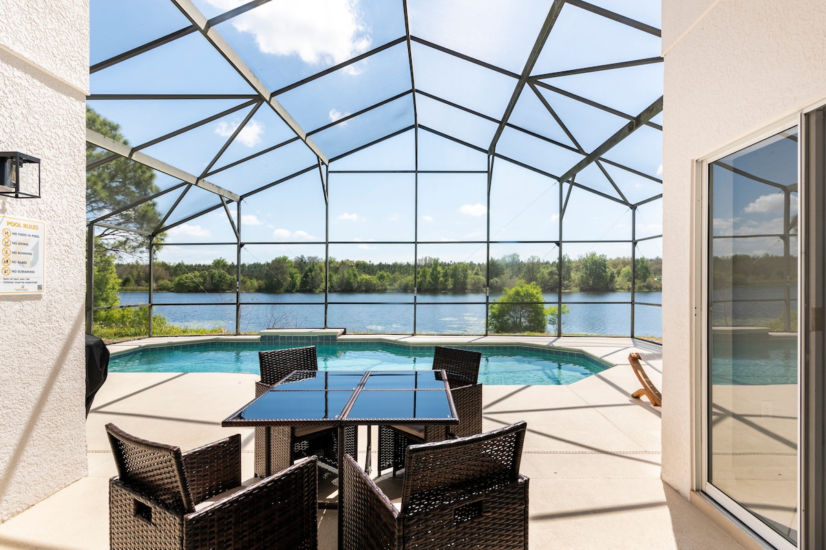 A private screened patio features a dining set with a glass table and wicker chairs, overlooking a serene lake. The clear blue sky is reflected in the water, while a pool glistens nearby, creating a peaceful outdoor space.