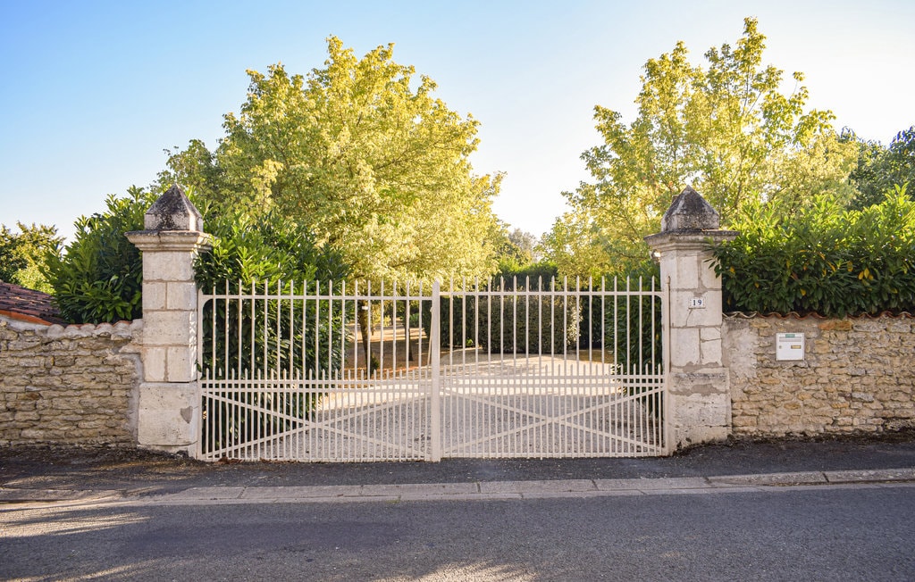 A gated entrance is framed by tall trees and lush greenery. The white wrought iron gates are set against a stone wall and lead into the expansive property. The road in front is paved, creating a welcoming approach to the surroundings.
