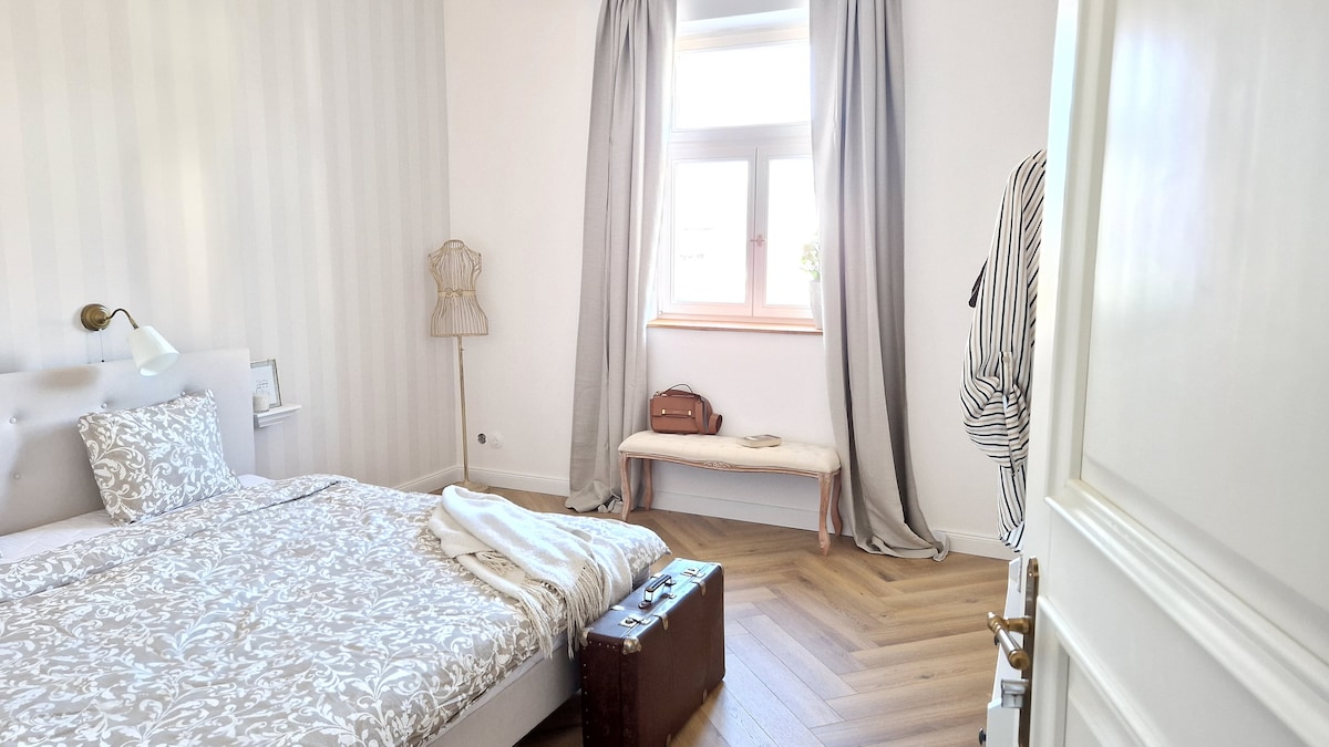 A serene bedroom features a spacious bed adorned with floral-patterned bedding. A window allows natural light to fill the room, while soft curtains frame the view. A bench and a vintage suitcase complement the minimalist decor, providing a welcoming and uncluttered atmosphere.