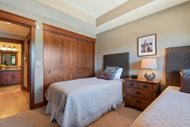 Un dormitorio organizado con dos camas individuales con cabeceras altas de tejido oscuro y cubrecamas texturizados de color gris claro, separadas por una mesita de noche de madera con tres cajones, una lámpara con base plateada y un despertador.