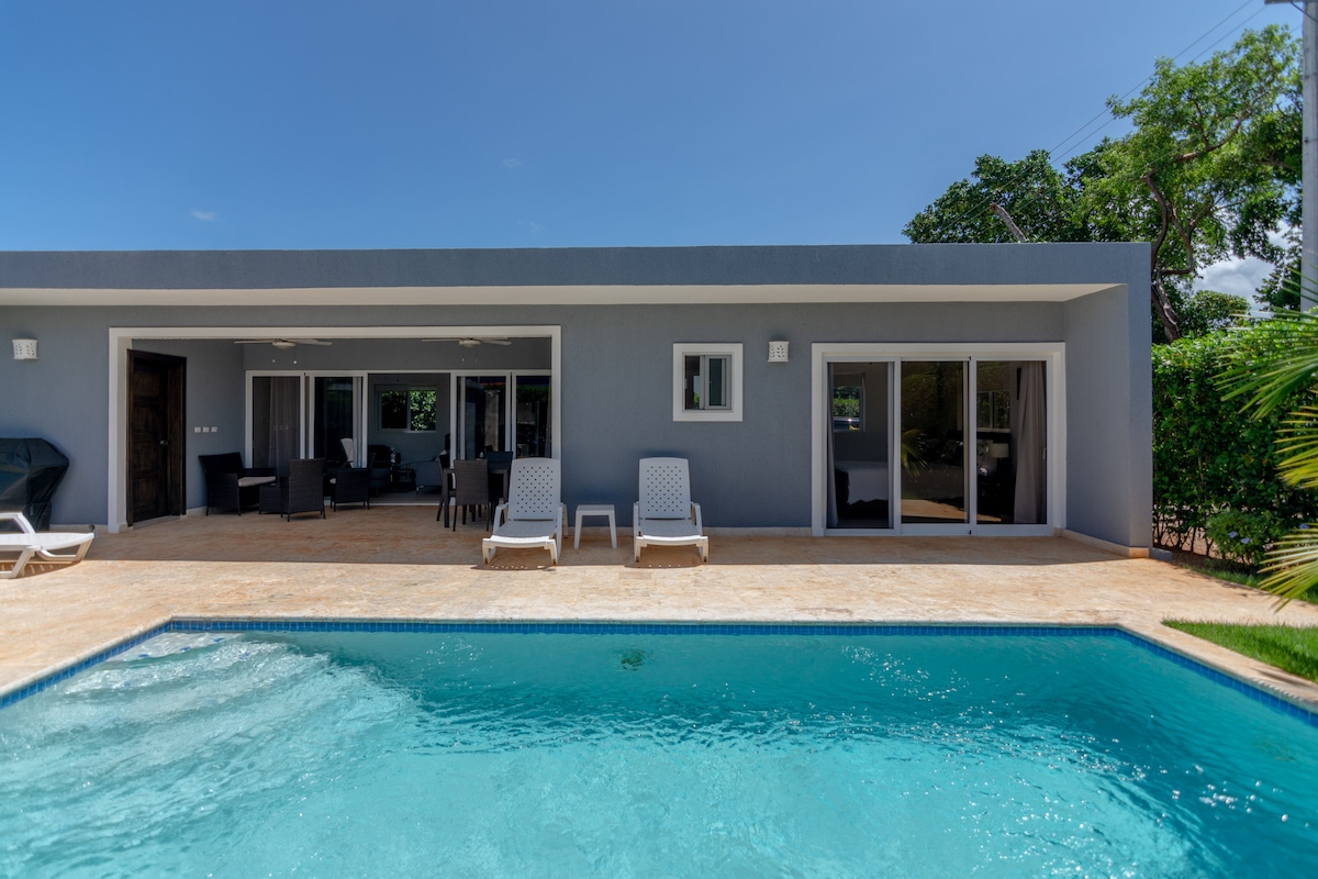 A modern villa features a private pool surrounded by a sun deck. Two lounge chairs sit next to the water. The expansive glass doors of the living area provide views of the outdoor space, highlighted by lush greenery in the background.