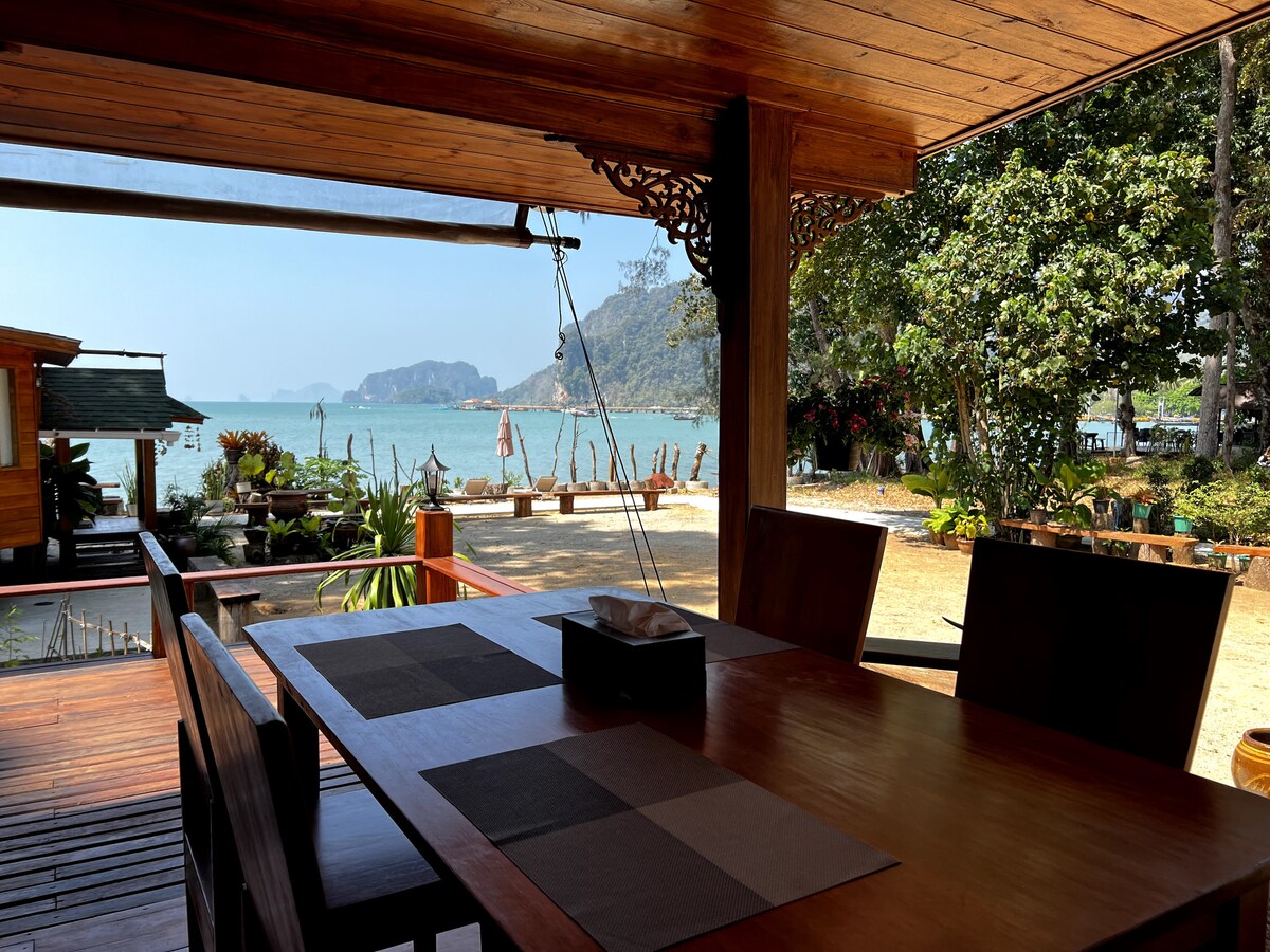 A dining area with wooden furniture overlooks a sandy beach and calm sea. The table is set with a placemat, while lush greenery can be seen in the background. Scenic mountains rise in the distance, enhancing the peaceful view.