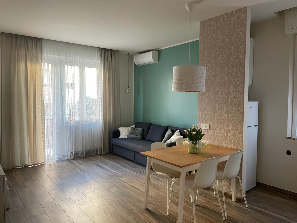A bright living area features a comfortable sofa, inviting natural light through large windows dressed in light sheer curtains. A small dining table with white chairs is positioned beside a patterned wall, and a refrigerator is visible in the kitchen area.