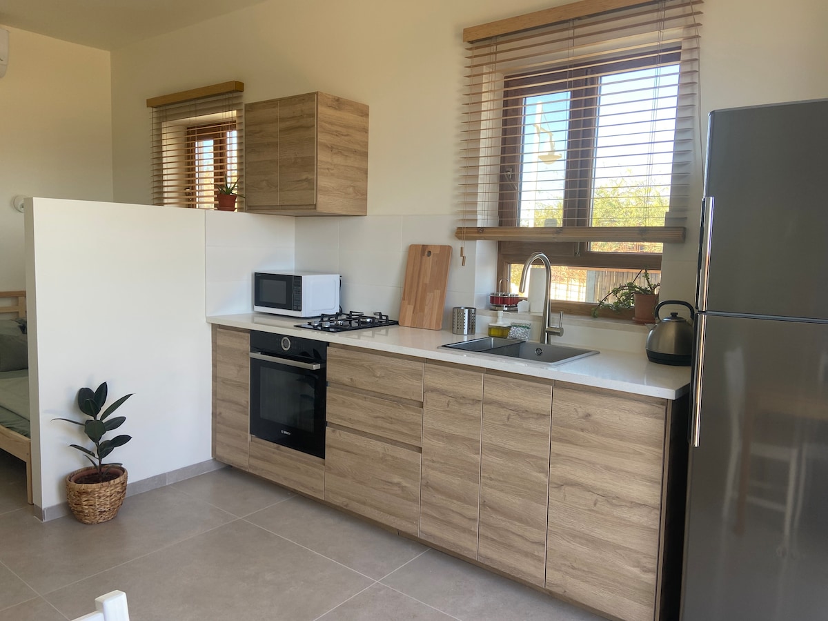 A modern kitchen is equipped with a stove, oven, microwave, and a refrigerator. Light wood cabinets and countertops complement the overall design. Natural light filters through the window blinds, illuminating plants placed on the ledge. A small potted plant adds a touch of greenery.