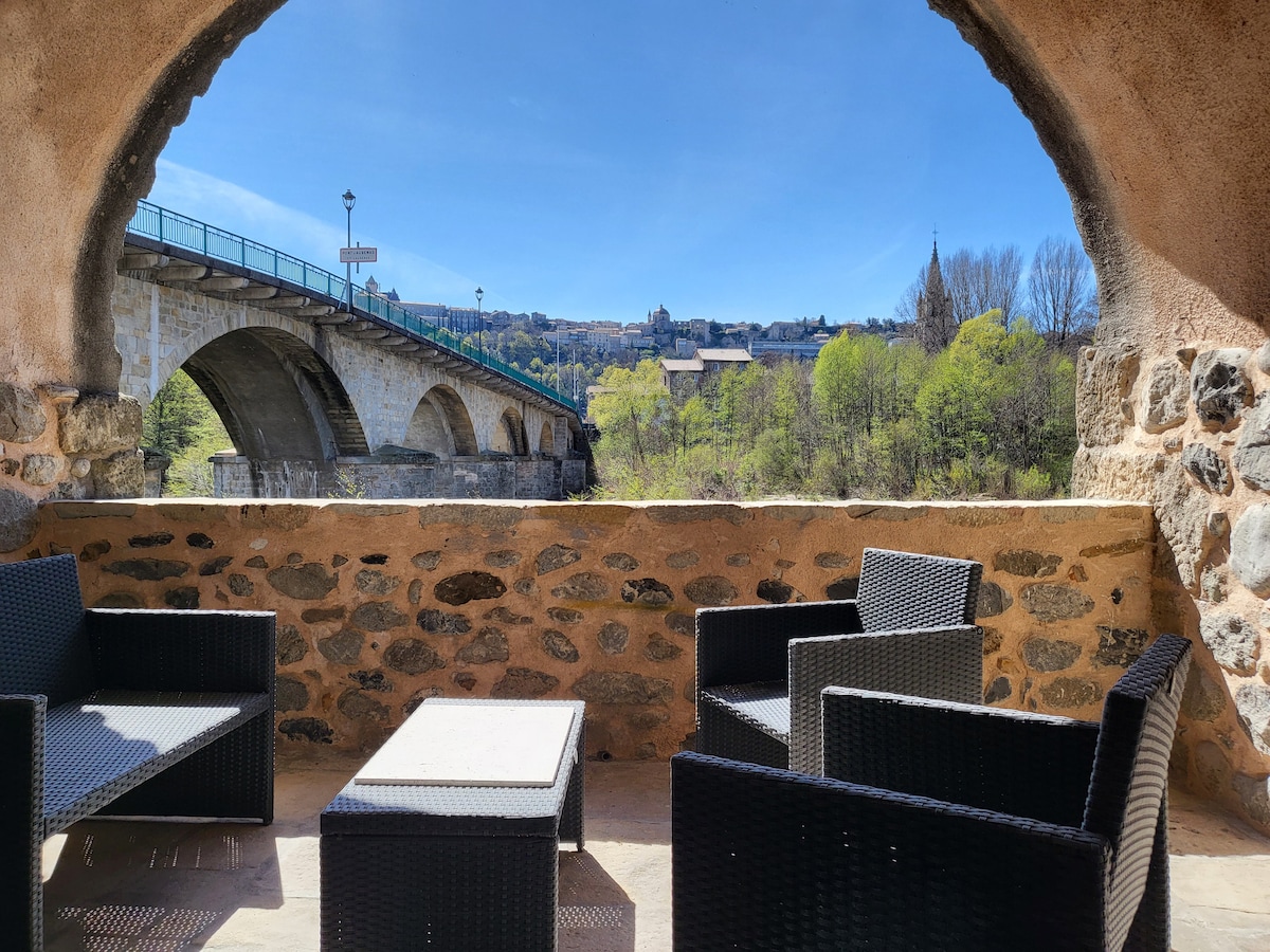 A cozy terrace is framed by a stone archway, featuring woven seating and a low table. The view beyond showcases a tranquil river and a bridge, with a backdrop of town and greenery under a clear blue sky.