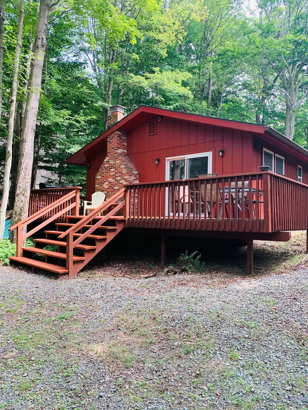 A rustic red cabin is framed by tall trees, featuring a wraparound deck with a set of stairs leading to the entrance. A stone chimney is visible on one side, and large windows provide views of the surrounding woods.
