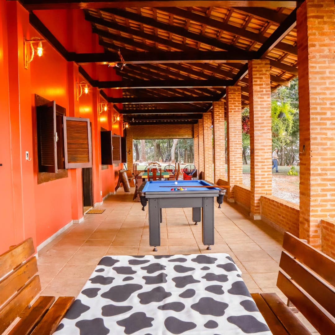 An open-air social area features wooden benches and a playful black-and-white patterned tablecloth. A pool table occupies the center, surrounded by warm red walls and exposed brick columns. Ample natural light filters through the windows, highlighting the inviting space.
