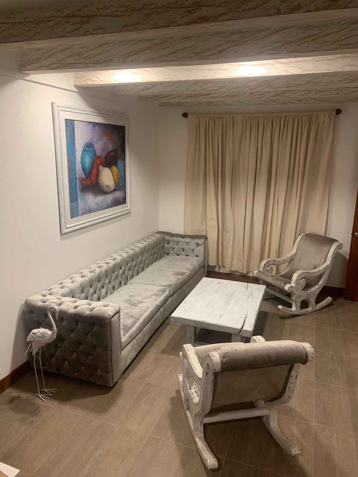 A comfortable seating area features a plush, tufted sofa and two armchairs. A white marble coffee table is centered in front of the seating. Soft curtains frame a window, and a colorful painting hangs above the sofa, adding visual interest to the neutral-toned space.