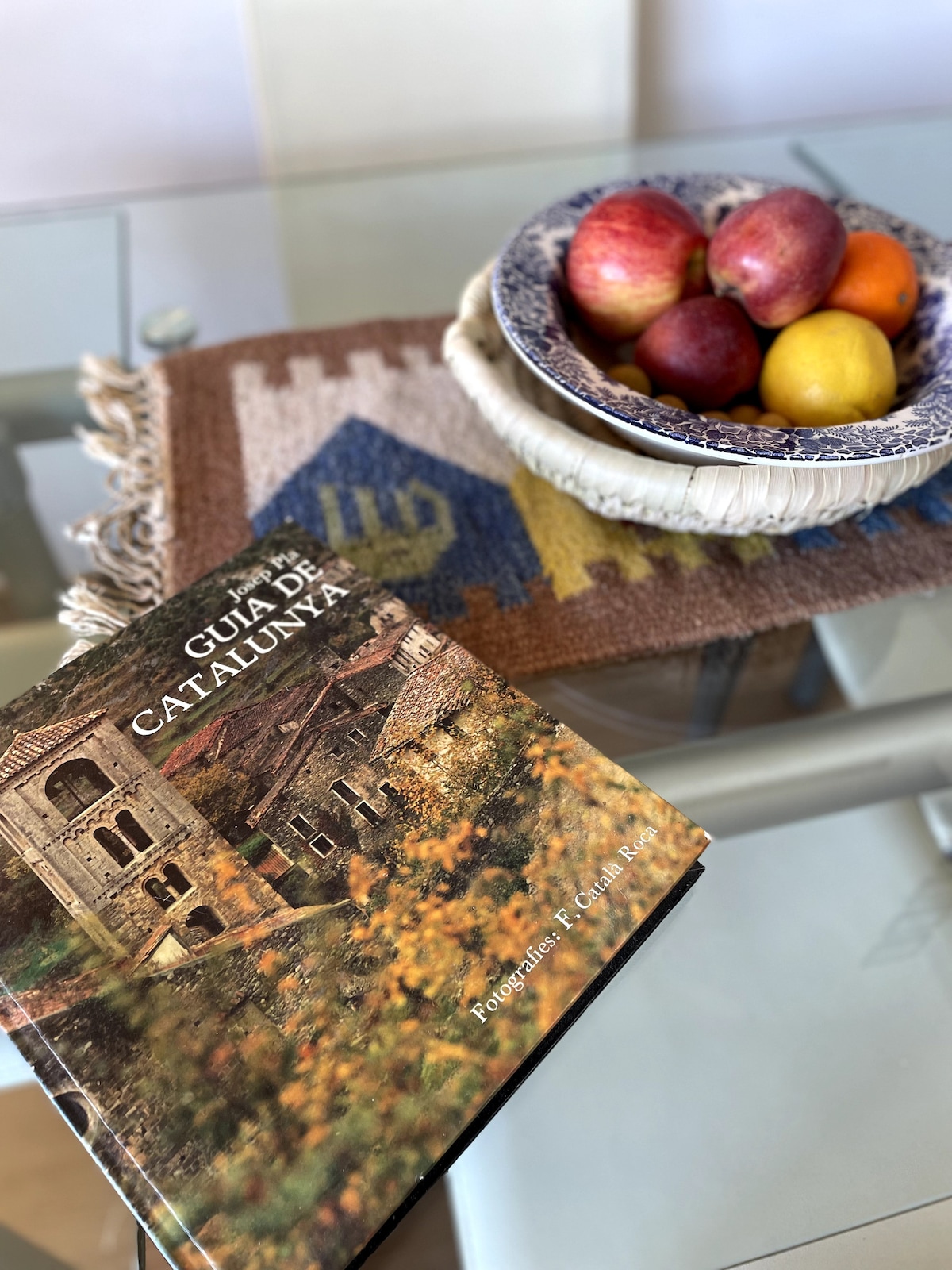 A neatly arranged dining table features a decorative woven placemat with a blue and yellow pattern. A bowl of red apples and yellow citrus sits alongside a guidebook titled 'Guia de Catalunya' with an image of a stone house surrounded by greenery.