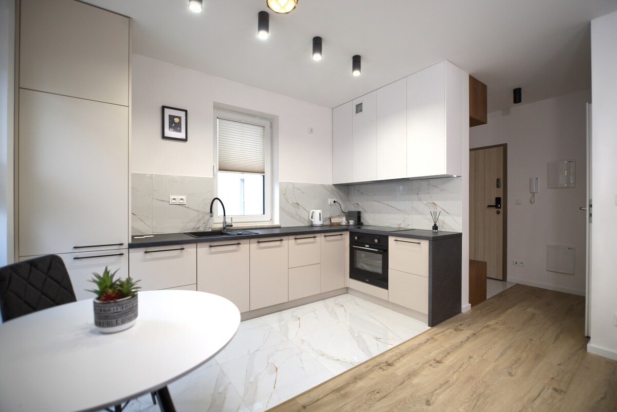 A modern kitchen is showcased with light-colored cabinetry and a dark countertop. Stainless steel appliances, including a microwave and oven, are integrated into the design. A round dining table with a plant centerpiece is positioned nearby, complementing the marble-patterned flooring.