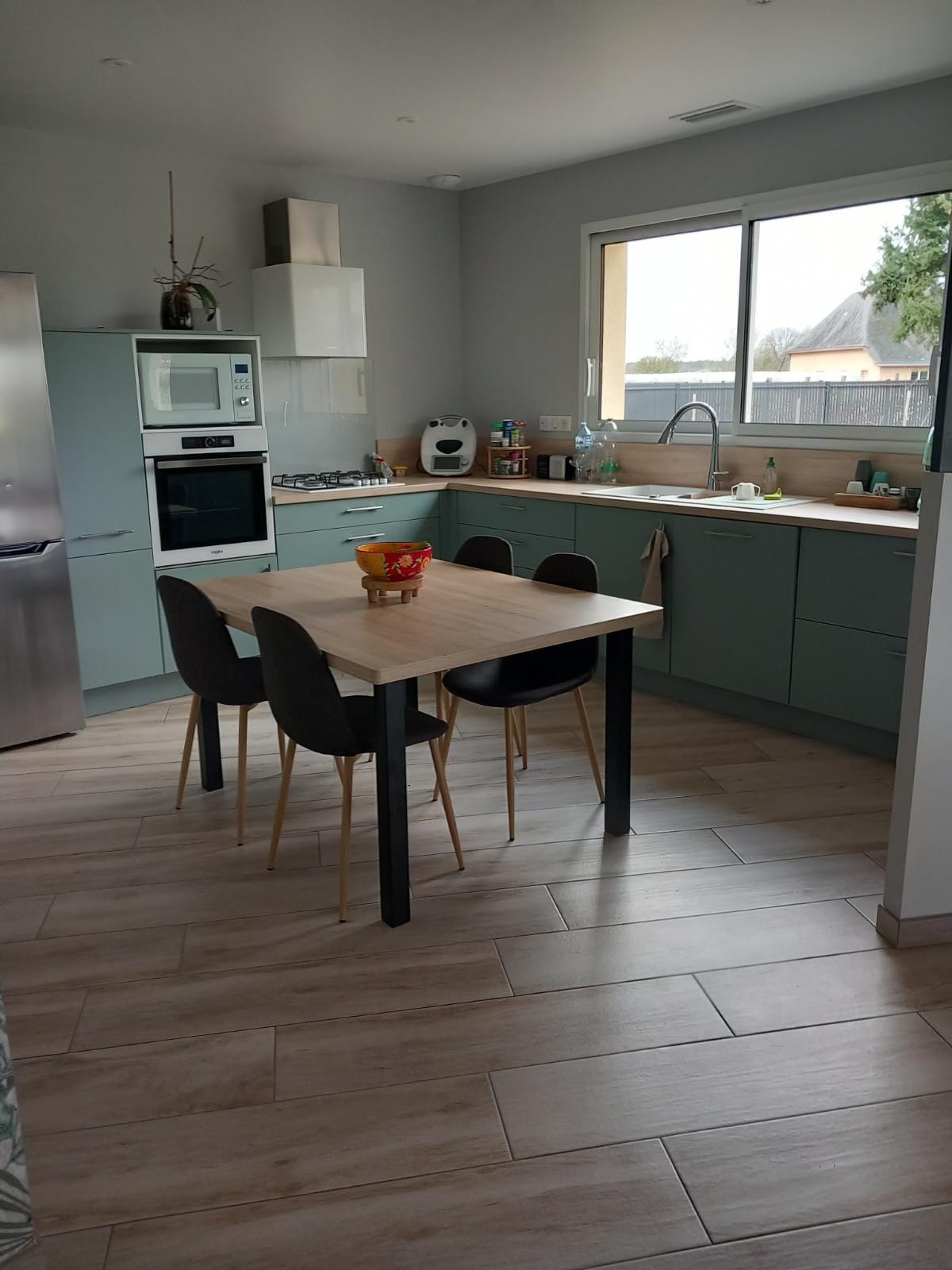 A modern kitchen features light green cabinets paired with a wooden table surrounded by four black chairs. Natural light fills the space through a large window above the sink, showcasing the stainless steel refrigerator and other appliances along the counter.