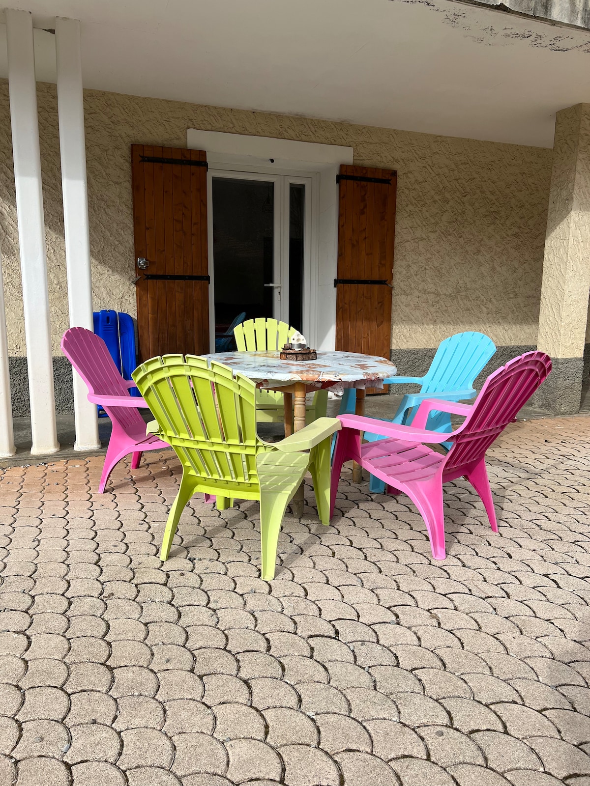 A colorful outdoor seating area is presented, featuring a round table surrounded by six Adirondack chairs in varying pastel shades of green, pink, and blue. The space is paved with stone tiles, and wooden doors are seen in the background, enhancing the inviting ambiance.