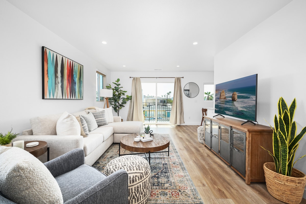 The living area features a comfortable sectional sofa paired with an accent chair and a decorative area rug. Large windows allow natural light to fill the space, while a flat-screen TV is positioned on a wooden console. A coffee table sits at the center, surrounded by greenery.