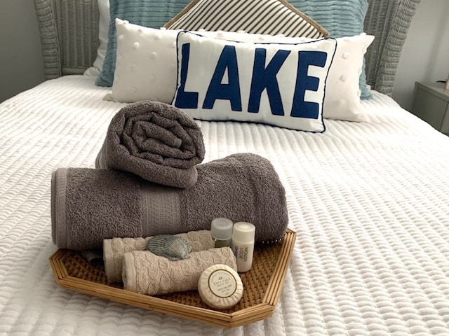 A neatly arranged tray is placed on a striped bedding set, featuring folded towels and natural bath products. A decorative pillow with the word 'LAKE' provides an inviting accent, while the overall setup suggests a relaxing environment.