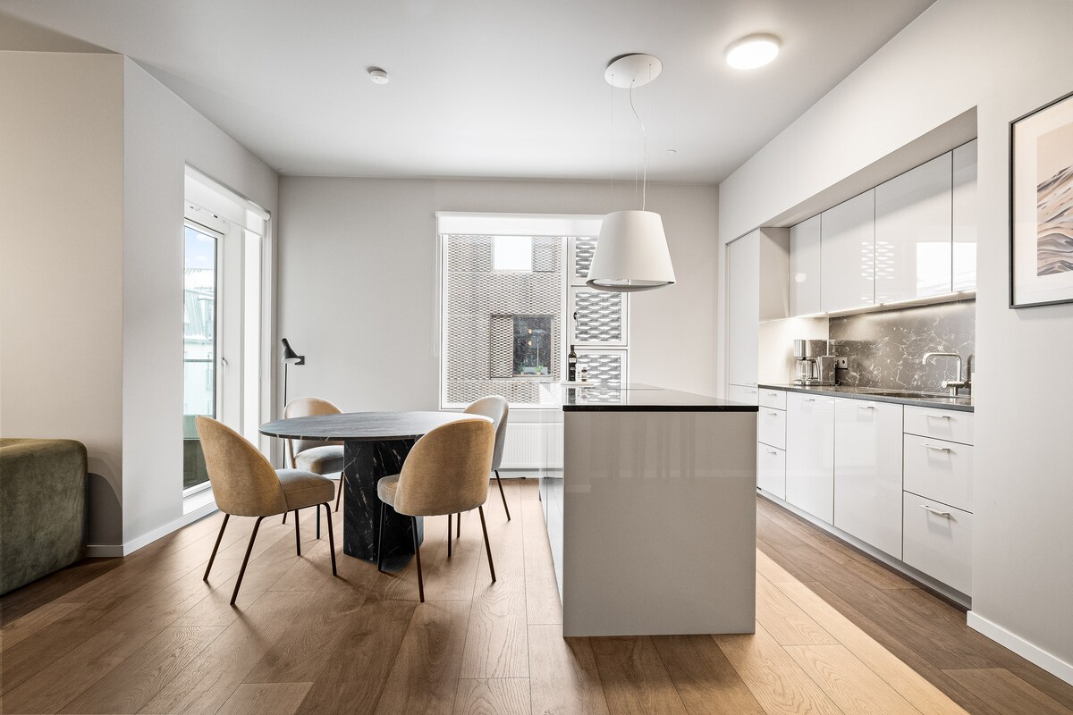 A modern kitchen and dining area is visible, featuring a sleek island with a dark countertop and light-colored cabinets. A round dining table with four upholstered chairs is positioned nearby, with natural light streaming in through large windows and glass doors.