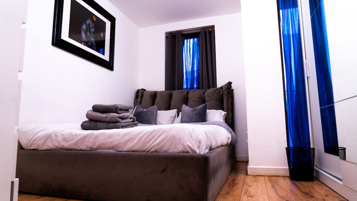 A spacious bedroom features a comfortable bed with layered bedding and decorative cushions. Soft blue curtains frame the window, allowing natural light to filter in. A framed artwork is displayed above the headboard, and a basket sits in the corner, complementing the minimalist decor.