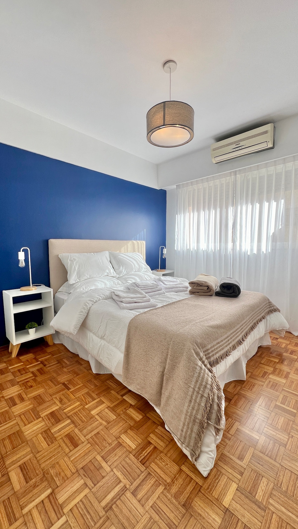 Chumba cha kulala kina kitanda chenye starehe cha ukubwa wa malkia kilicho na matandiko ya kifahari, ukuta wa rangi ya bluu na mwanga wa kutosha wa asili kutoka kwenye madirisha makubwa. Meza mbili za kisasa kando ya kitanda zilizo na taa na taa nzuri ya juu hukamilisha mazingira ya kuvutia na maridadi.