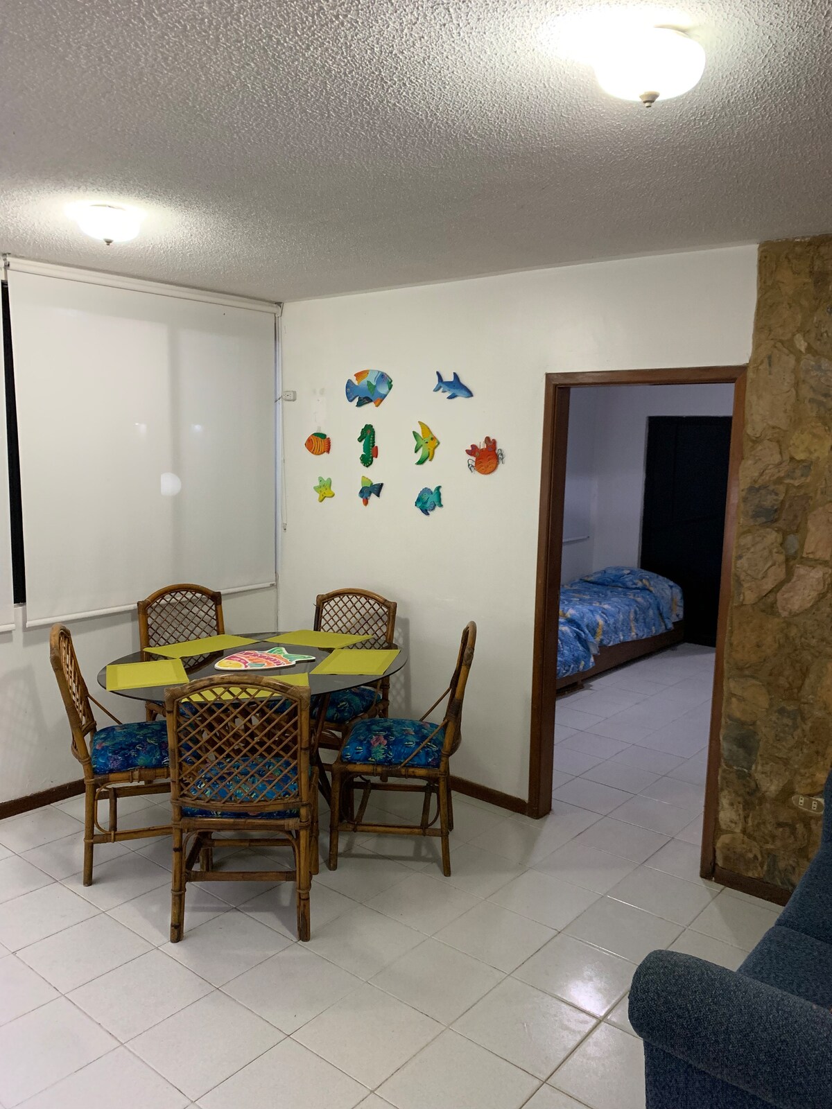 A dining area features a round table surrounded by four woven chairs, with colorful fish decorations adorning the wall. Natural light enters through a large window covered by light shades. An arched doorway leads into a bedroom with a bed covered in blue bedding.