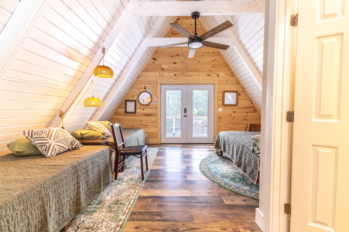 An open sleeping area is framed by sloped ceilings and wooden walls. Two twin beds are dressed with dark green covers, while decorative pillows add visual interest. A chair is positioned beside a small area rug, and large glass doors lead to an outdoor deck.