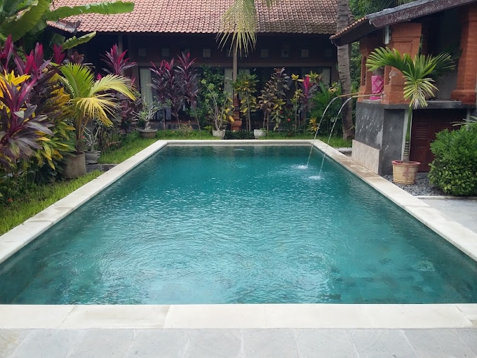 A shared swimming pool is surrounded by lush tropical plants and colorful foliage. Sunlight reflects off the clear water, which features a gentle fountain at one end. The serene setting is enhanced by a well-maintained garden area, offering a peaceful retreat for guests.