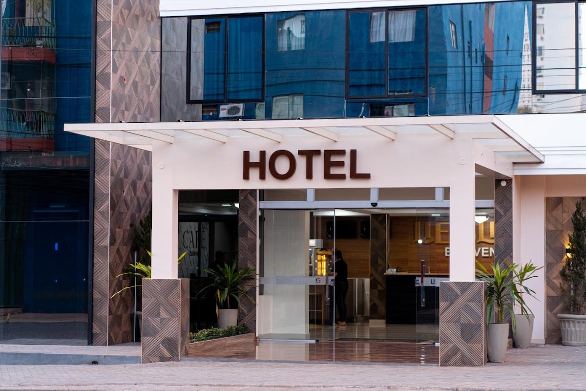 The hotel's entrance is characterized by a modern design, featuring a large sign that reads 'HOTEL.' Glass doors provide a clear view of the reception area, while potted plants add a touch of greenery to the welcoming facade.