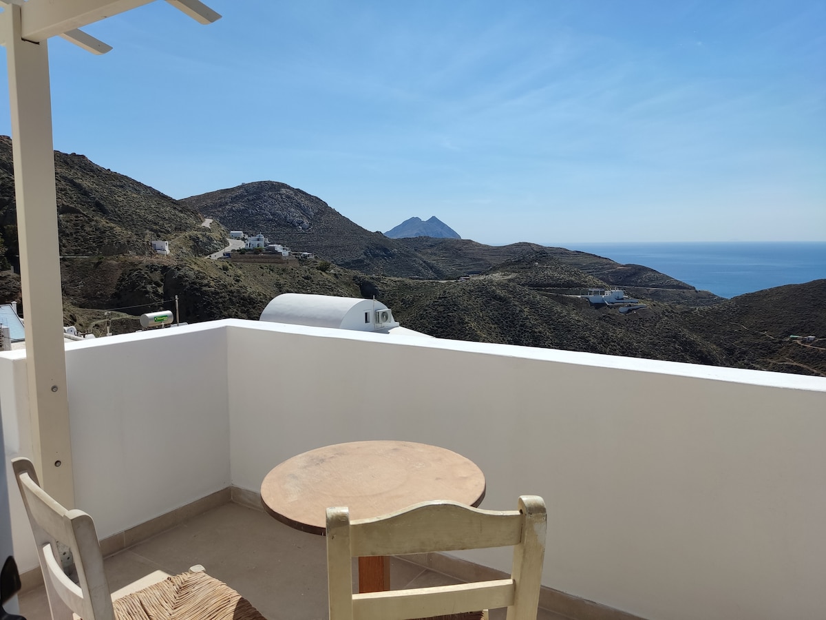 A simple wooden table and chair are placed on a private balcony, overlooking a scenic mountain landscape and the coastline. The clear sky adds brightness to the view, while the distant mountains are visible across the sea.