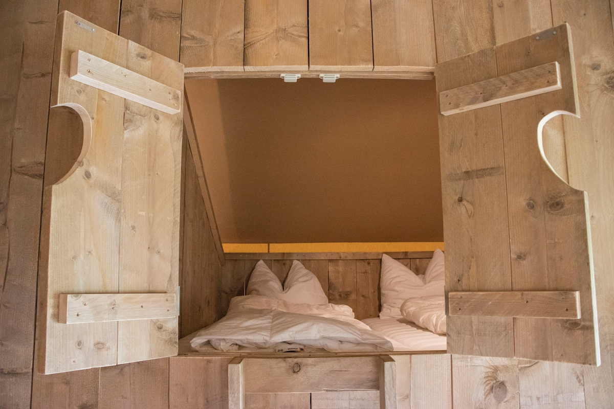 Lofted sleeping area is framed by rustic wooden walls, featuring two cozy beds tucked beneath a softly sloped ceiling. The wooden shutters are opened, allowing natural light to filter in, enhancing the warm and welcoming ambience of the space.