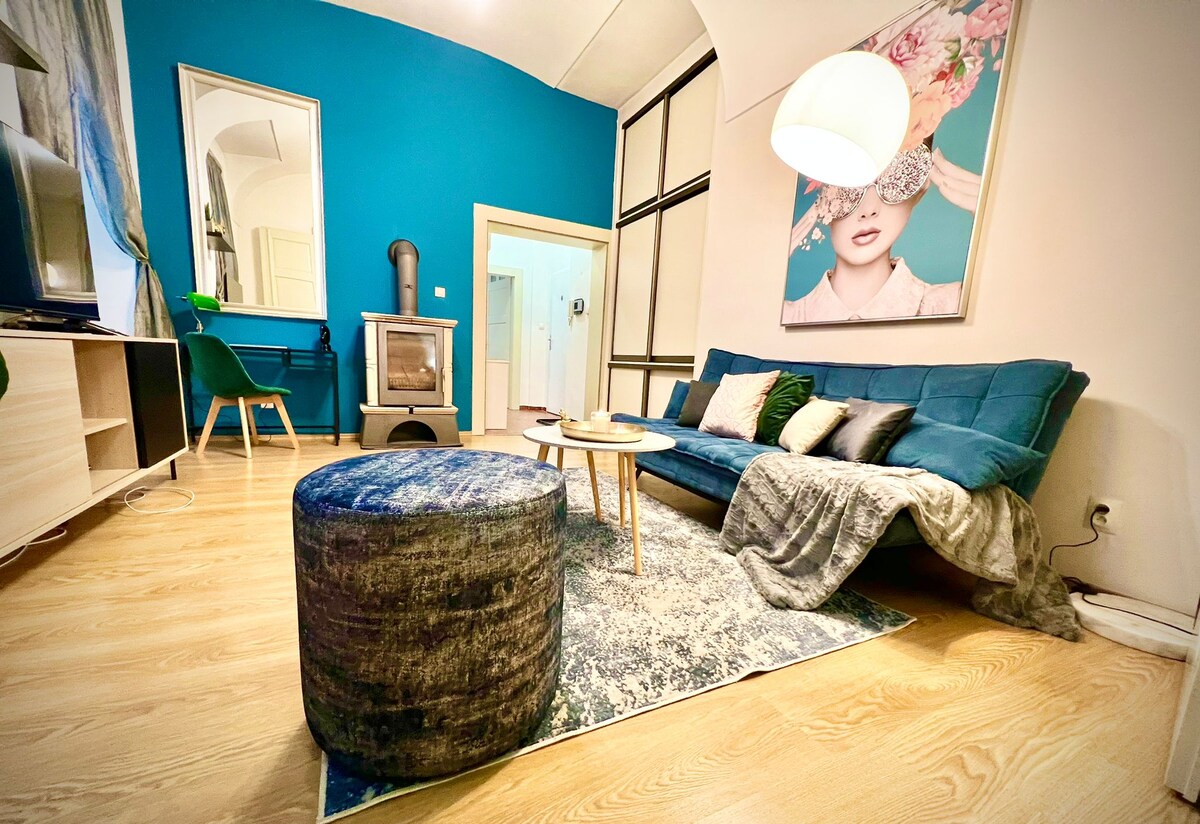 A cozy living area features a blue velvet sofa paired with a soft, textured ottoman and a round table. A warm wood floor complements the space, while a modern lamp provides overhead lighting. A decorative rug anchors the seating area, enhancing the stylish decor.