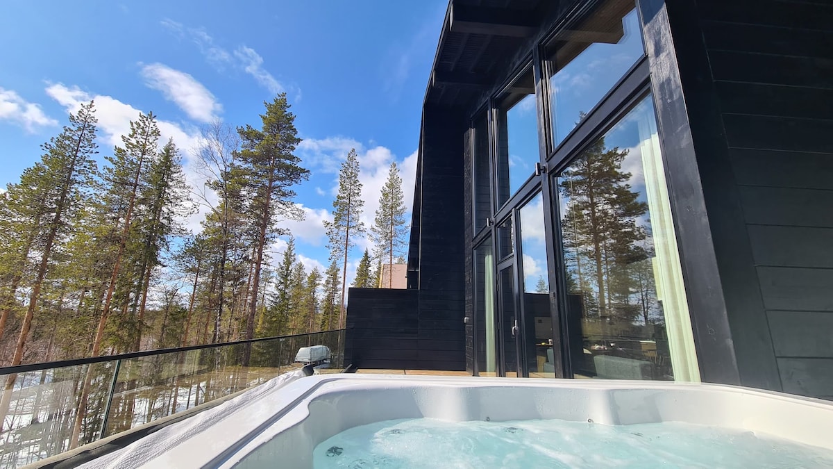 An outdoor hot tub is positioned on a spacious terrace, offering a view of tall trees and a clear blue sky. The villa's modern design features large windows that enhance the connection to nature. The tranquil setting invites relaxation and enjoyment of the surrounding landscape.