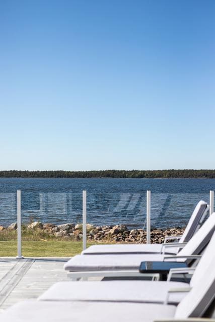 A deck area features several loungers facing a tranquil body of water. Glass panels provide a clear view of the landscape, which includes a shoreline lined with rocks and a backdrop of lush greenery under a clear blue sky.