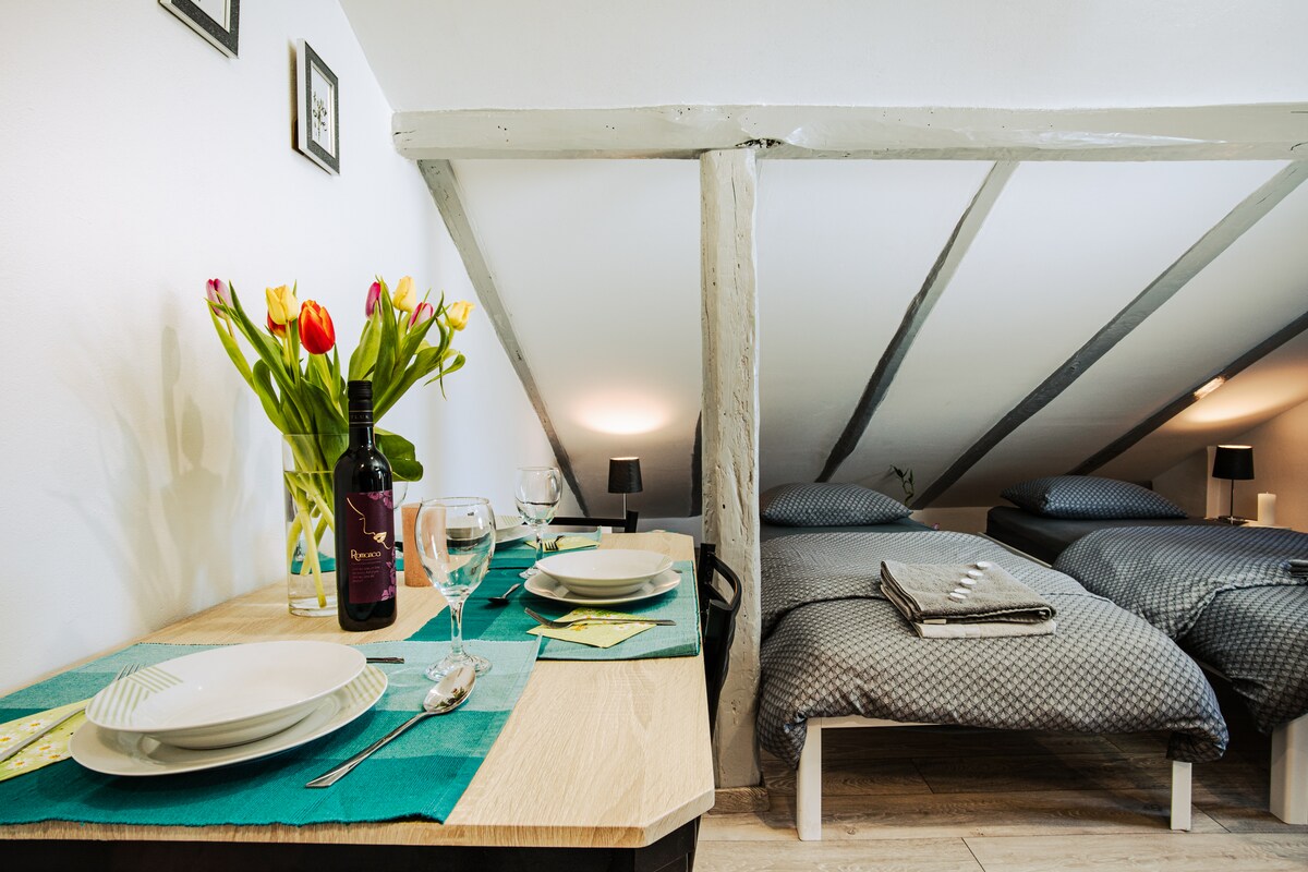 A dining area features a wooden table set with plates and glasses, accompanied by a bouquet of tulips. In the background, two neatly made beds with gray patterned bedding are visible under sloped ceiling beams, which add character to the space.