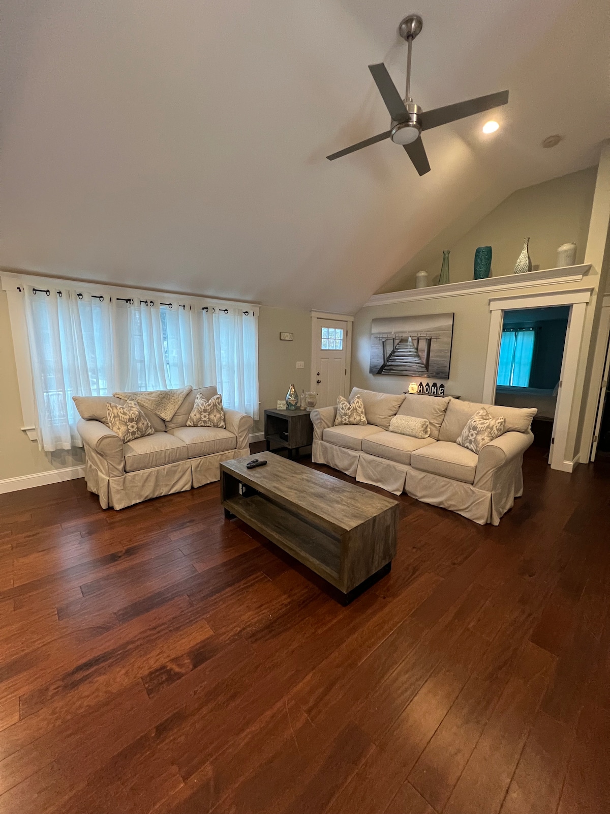 A warm living area features two cushioned sofas adorned with decorative pillows, facing a sturdy wooden coffee table. Natural light filters in through multiple windows lined with sheer drapes, highlighting the rich hardwood flooring. Subtle decorative accents adorn the shelves in the background.