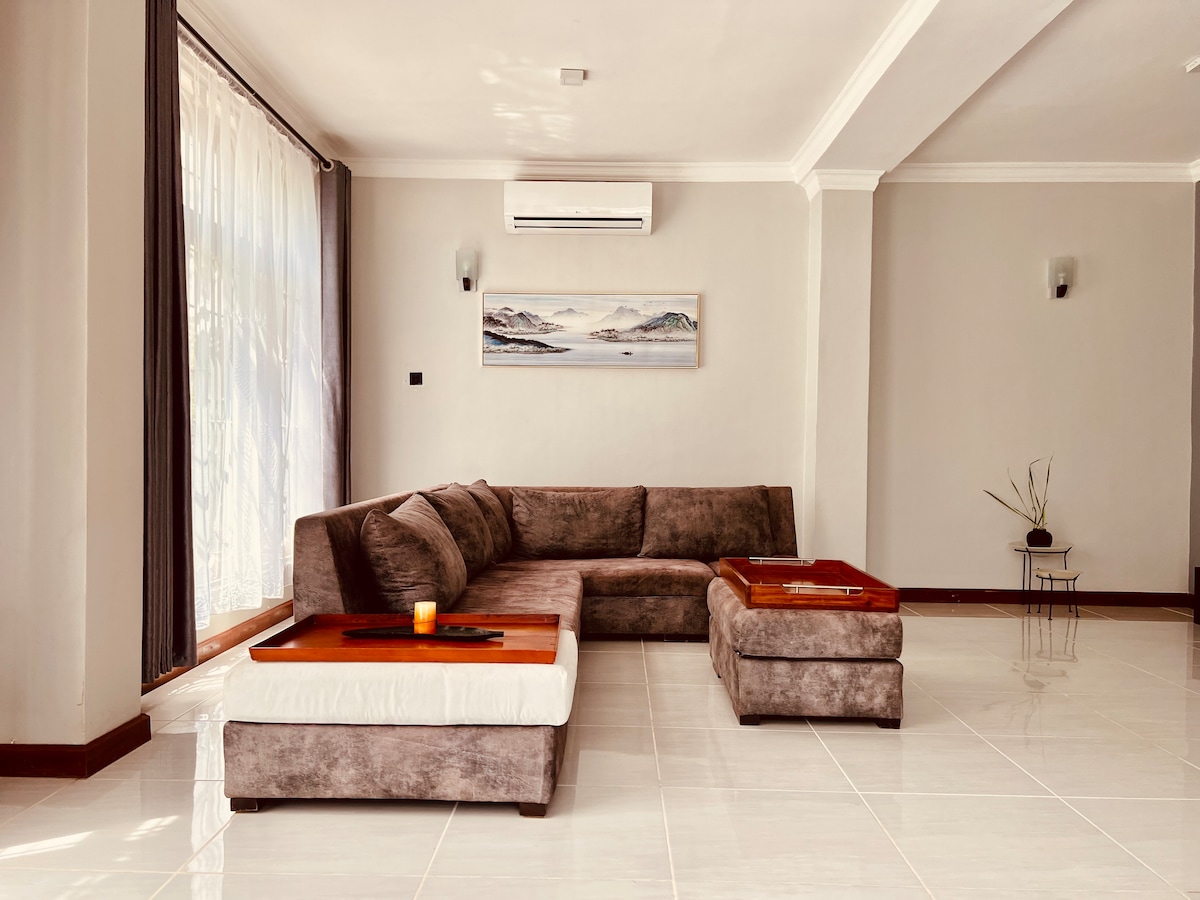 A spacious living area features a plush sectional sofa with soft cushions. Two decorative wooden tables are positioned in front, enhancing comfort. Natural light streams through large windows, with sheer curtains softly diffusing the sunlight. Minimalist decor adds to the tranquil environment.
