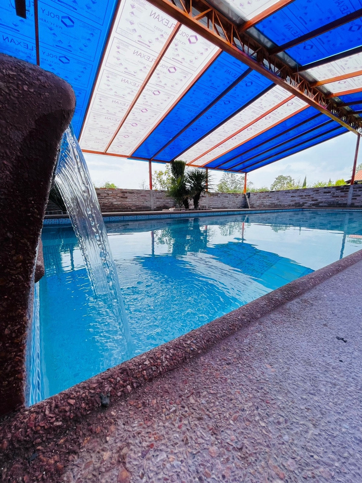 A serene swimming pool is framed by a textured stone wall, with water cascading from a decorative feature. A colorful cover provides shade, while the blue water reflects the vibrant hues above, creating a peaceful atmosphere.