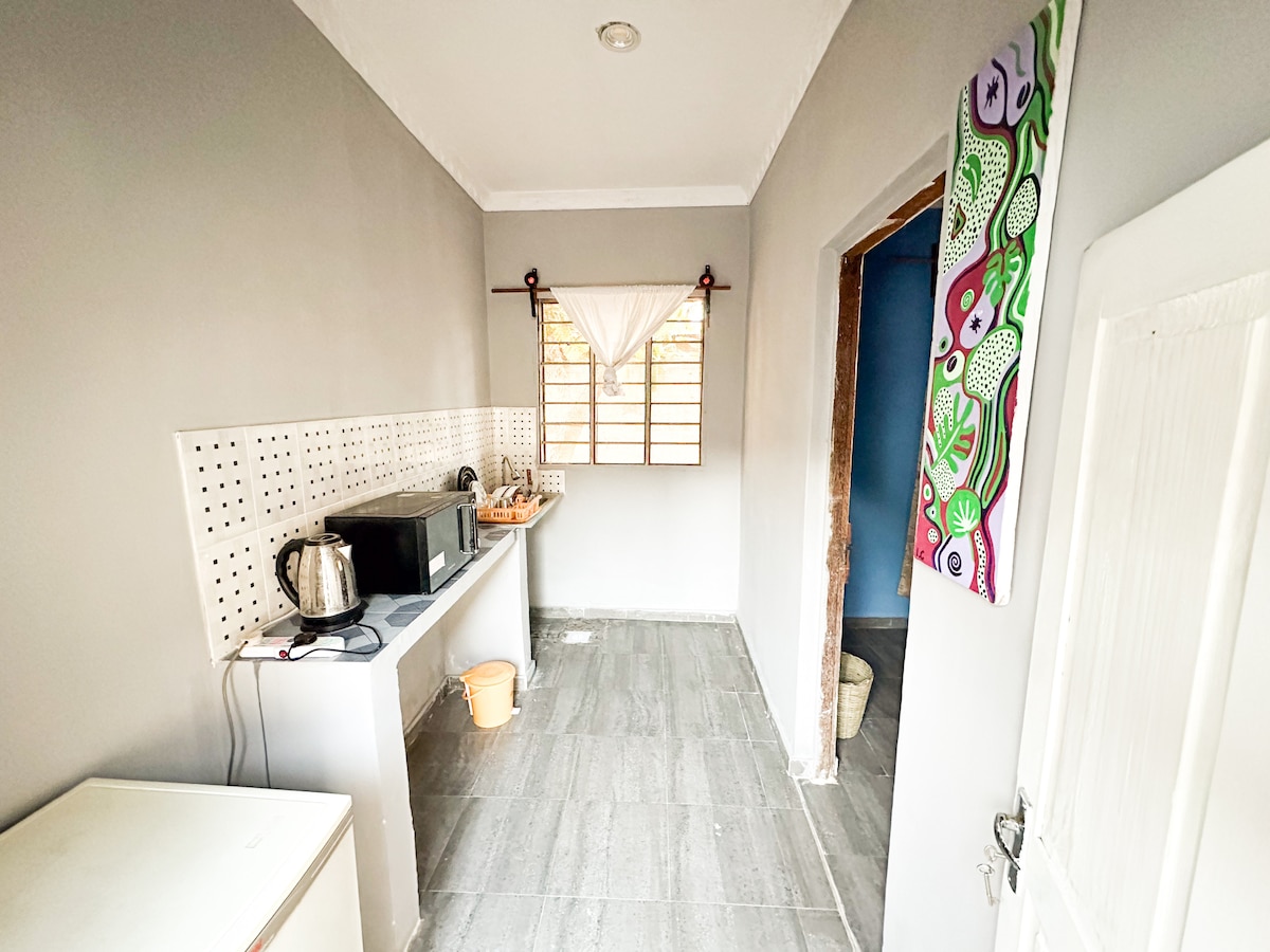 A functional kitchen area is presented with sleek gray walls and tiled flooring. Modern appliances, including a kettle and microwave, are available on the countertop. Natural light enters through a window adorned with a light curtain, and a colorful piece of wall art adds a subtle decorative touch.
