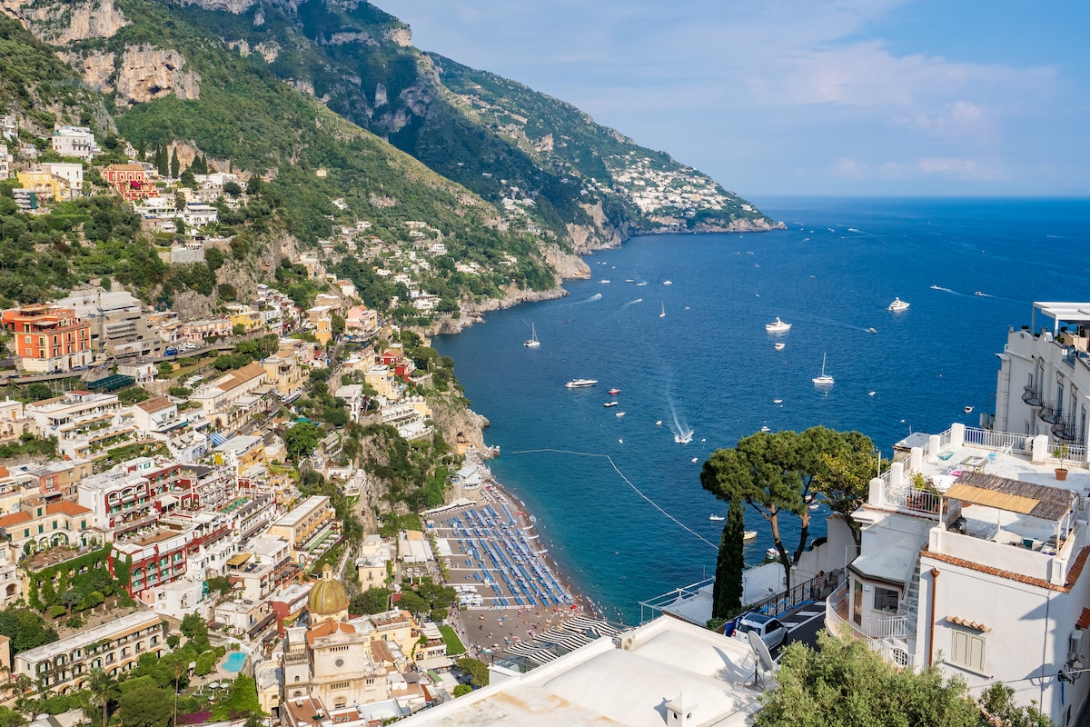 A breathtaking view captures the coastline of Positano, featuring colorful buildings cascading down the hillside towards the beach and the azure sea. Sailboats dot the water, while vibrant beach umbrellas line the shore, framed by lush greenery and rocky cliffs in the background.
