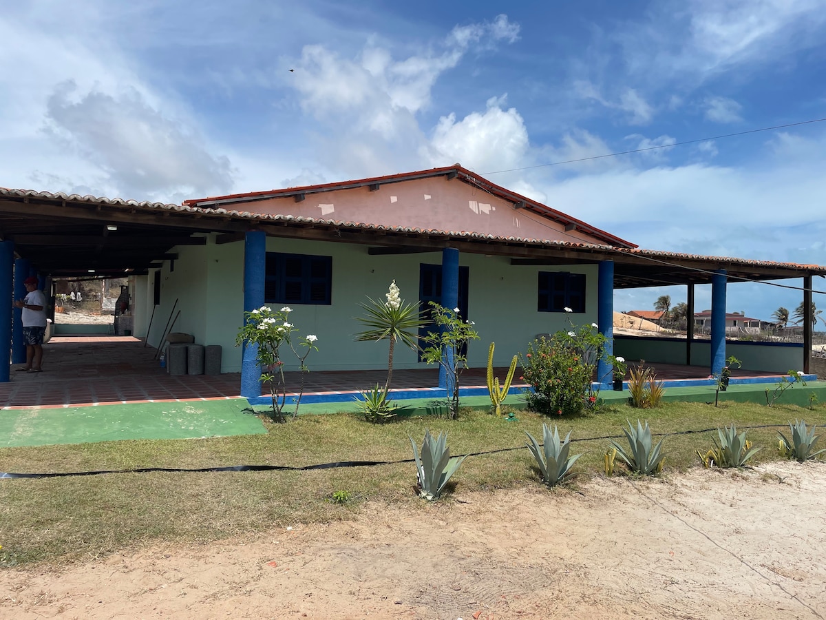 A simple coastal house is depicted with a blue and green exterior. A covered porch stretches across the front, providing ample shade. Tropical plants and flowers line the walkway, leading to a sandy area. The sky is clear, suggesting a pleasant outdoor environment.