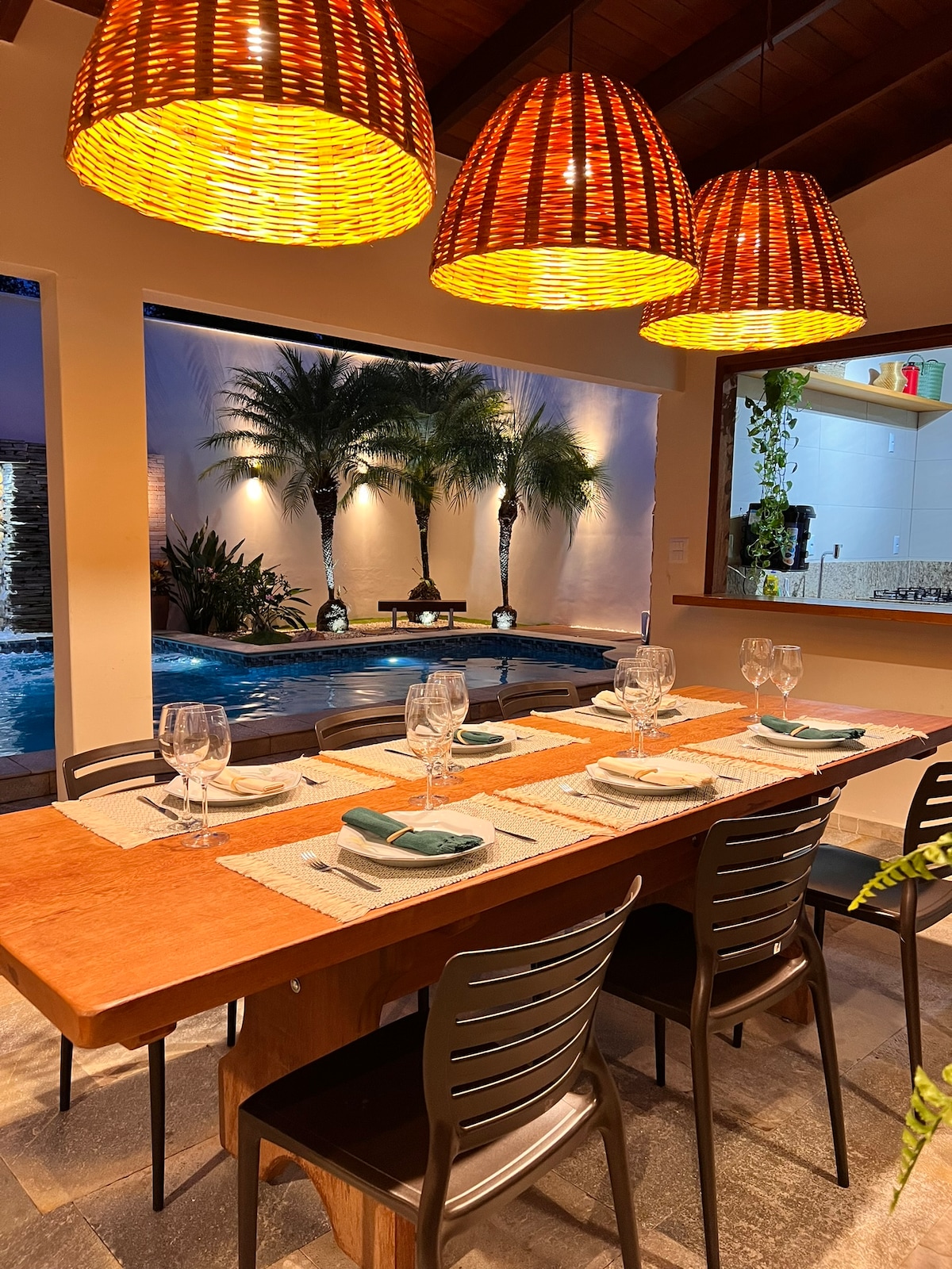 A dining area is presented with a wooden table set for six, featuring elegant glassware and neatly arranged placemats. A backdrop of lush palms is visible through large windows, alongside a glistening pool illuminated by ambient lighting. Unique woven pendant lights hang overhead.