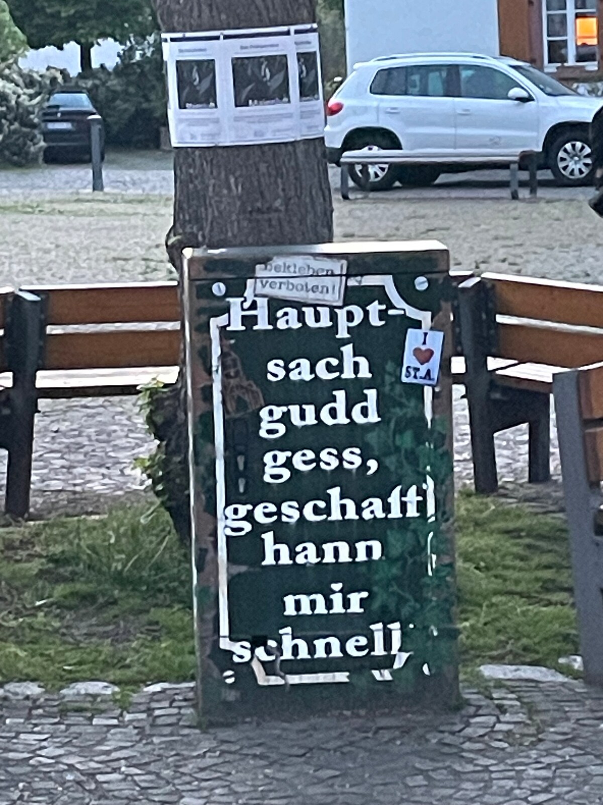 A wooden sign at a park is adorned with bold white lettering, featuring a message in a local dialect. Surrounding seating includes benches under a large tree, contributing to a serene environment. A notice can be seen attached to the tree in the background.
