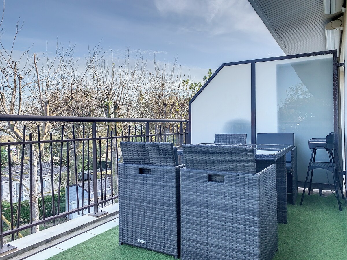 A terrace area features a set of dark wicker chairs and a dining table, surrounded by a railing. Lush greenery and bare trees are visible beyond, with a clear sky completing the outdoor scene.
