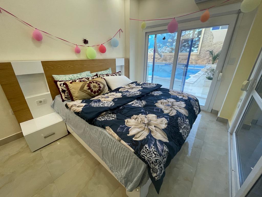 A comfortable bedroom features a spacious bed adorned with decorative pillows and a patterned blanket. Large glass doors lead to the pool area, allowing natural light to fill the room. Colorful decor elements are present along the wall, adding a cheerful touch.