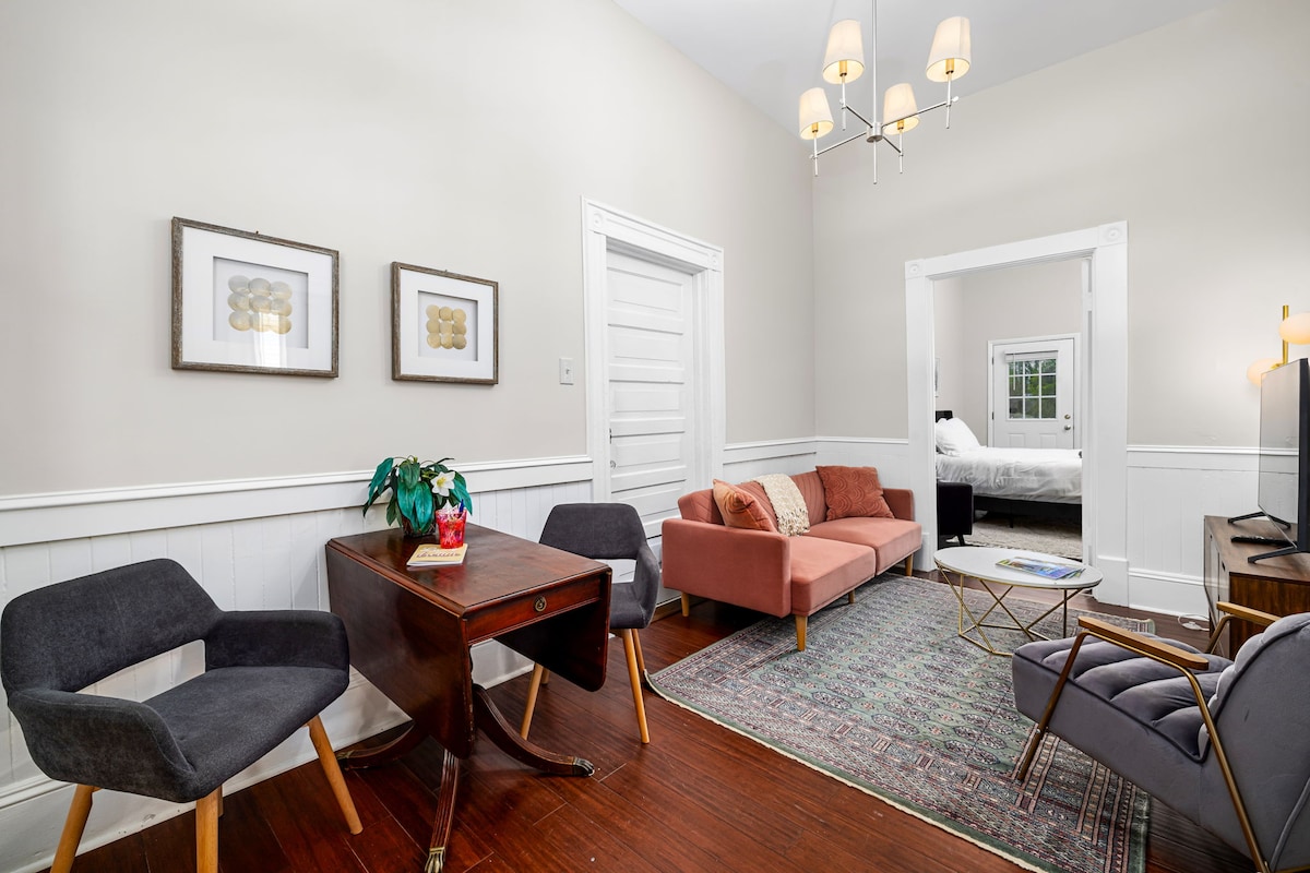 A common area is showcased with a vibrant rug and seating options including two chairs and a pull-out couch. A wooden desk is positioned nearby. Framed artwork decorates the walls, and a TV stands on a stand. A doorway leads to a bedroom in the background.