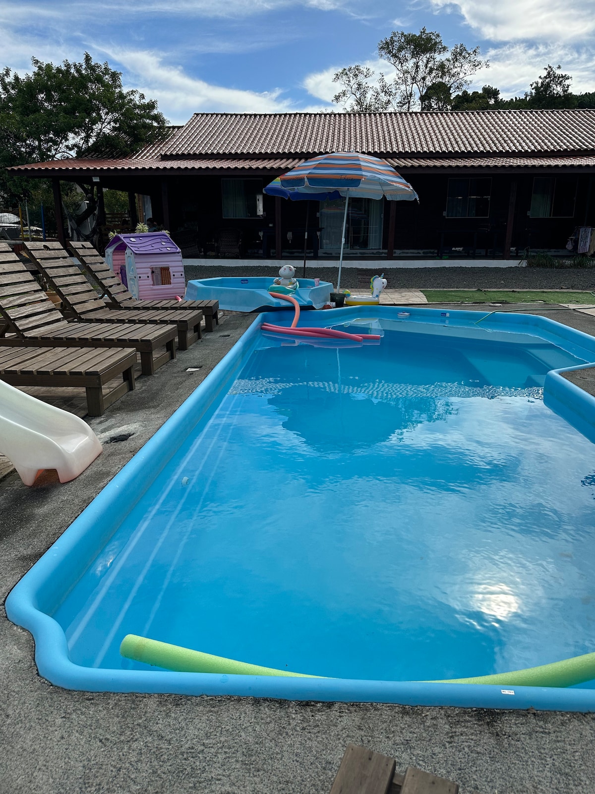 A clear swimming pool is framed by a shaded area with lounge chairs and an umbrella. Colorful pool toys float on the water's surface. A children's playhouse is positioned nearby, adding a playful touch to the outdoor space.