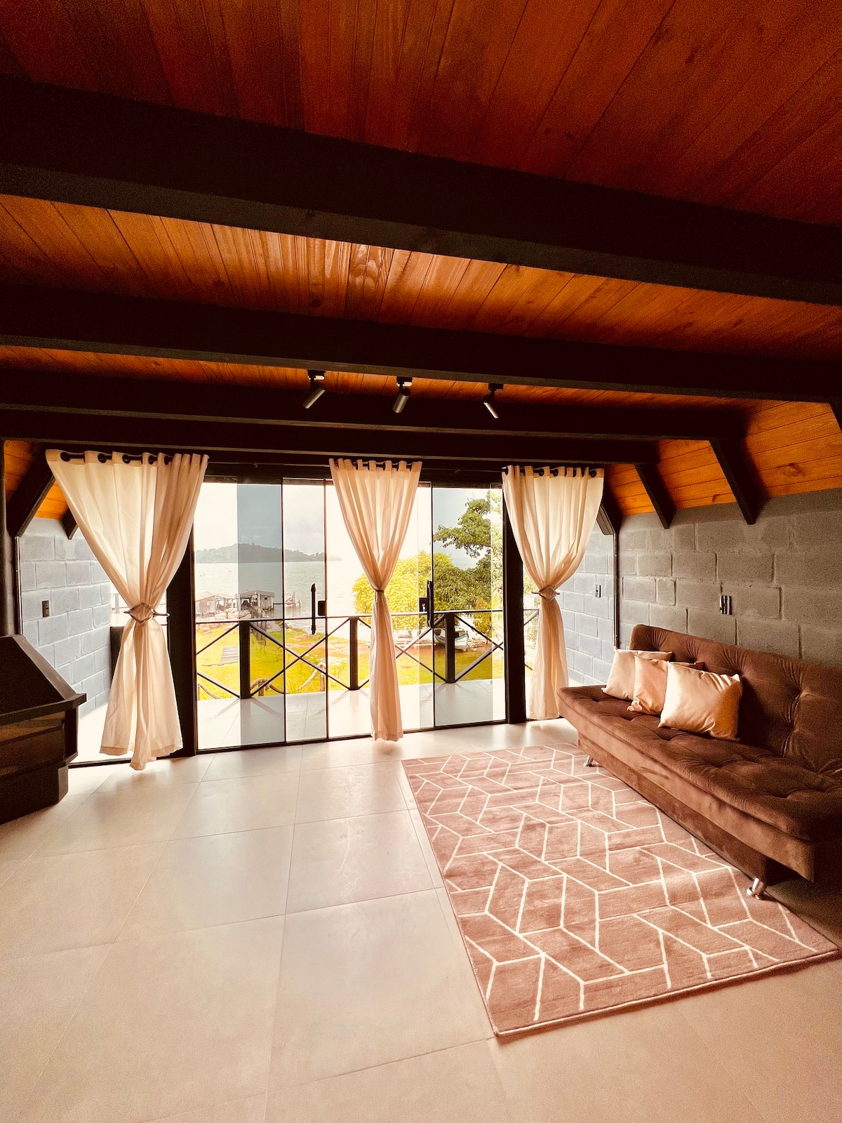 A spacious living area features wood-paneled ceilings and large glass doors that open out to a scenic view. Soft curtains frame the windows, allowing natural light to fill the space. A cozy sofa is positioned across from the doors on a geometric-patterned rug.